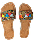 A pair of tan leather slide sandals with colorful geometric beaded straps, displayed on a brown textured fabric background.