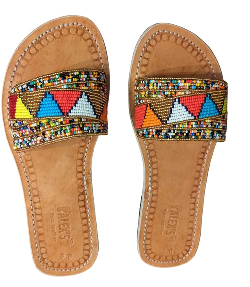 A pair of tan leather slide sandals with colorful geometric beaded straps, displayed on a brown textured fabric background.